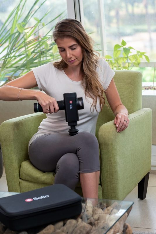 Woman applying Solio Super Gun Massager on leg while sitting